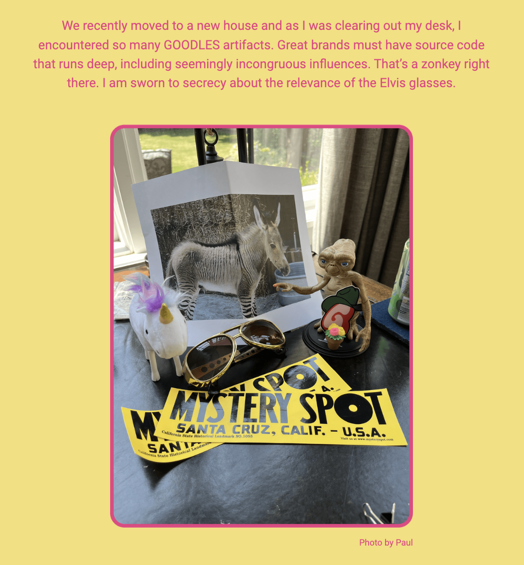 The image features a variety of items arranged on a surface, with a background text overlay. Here is the alt text including all of the text that appears in the image:

"A collection of quirky items on a desk, including a photograph of a 'zonkey' (zebra-donkey hybrid), a figurine resembling a famous movie alien in a red shirt holding a pizza, a plush unicorn toy, and a pair of oversized sunglasses. In the foreground, there are two yellow tickets with 'MYSTERY SPOT SANTA CRUZ, CALIF. - U.S.A.' printed on them. The overlaid text reads: 'We recently moved to a new house and as I was clearing out my desk, I encountered so many GOODLES artifacts. Great brands must have source code that runs deep, including seemingly incongruous influences. That’s a zonkey right there. I am sworn to secrecy about the relevance of the Elvis glasses. Photo by Paul'."