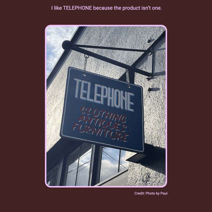 The image features a hanging sign with the word "TELEPHONE" prominently displayed, with additional text underneath. Here is the alt text including all of the text that appears in the image:

"A square hanging sign with a dark background and the word 'TELEPHONE' in large, cut-out letters at the top, under which 'CLOTHING ANTIQUES FURNITURE' is written in smaller letters. The sign is suspended from a metal bracket and is pictured against a textured building façade with a window reflecting the sky. Above the image, the text reads: 'I like TELEPHONE because the product isn’t one.' The image has a pink border, and at the bottom right corner, it is credited with 'Photo by Paul'."
